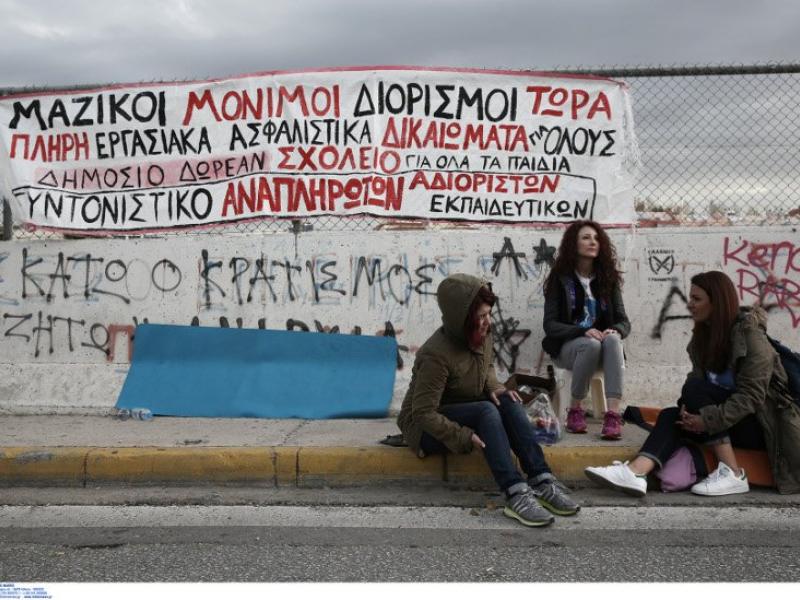 Εκπαιδευτικοί κάθονται σε πεζοδρόμιο με πανό από πίσω τους